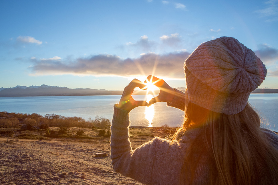 Girl holds hands in the shape of a heart in front of sunset.